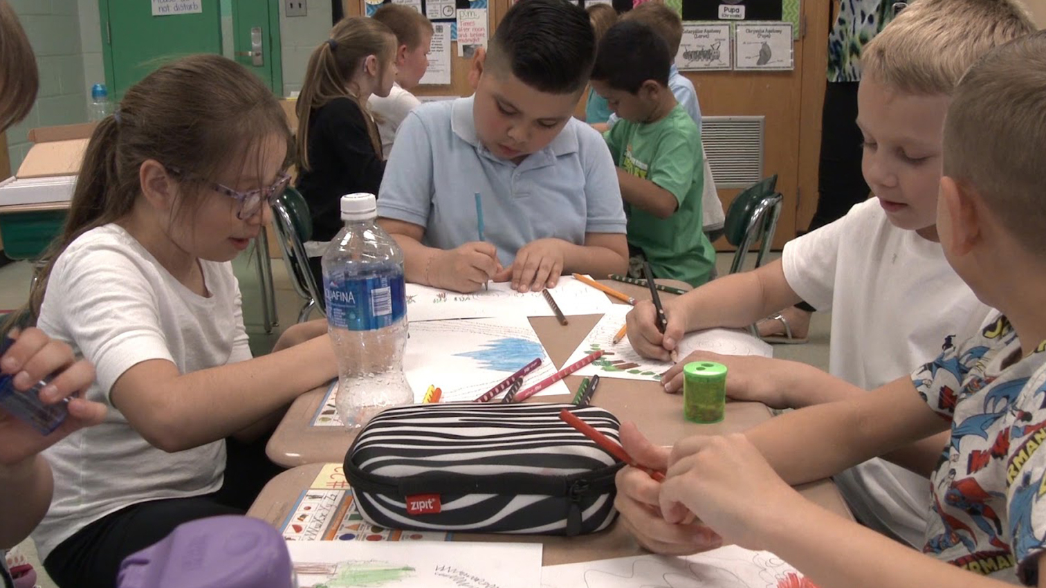 Group of students seated at desks working on drawn settings with colored pencils.