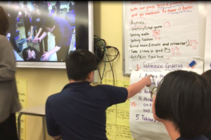 Back of a middle school student's head as he points to a chart labeled "Tableaux Criteria." To his left is a smartboard with a paused video of fellow students acting a scene.