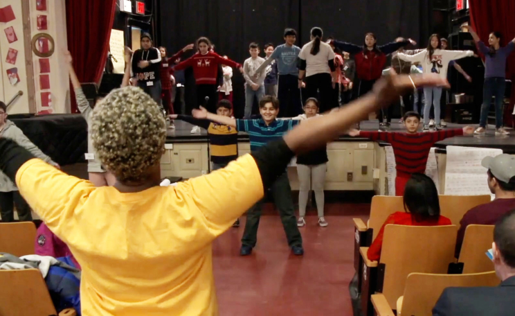 Students take a dance class in a school auditorium. The dance teacher has her back to the camera, she raises her arms in a warm-up and a class of middle school students facing her mirror her movement.