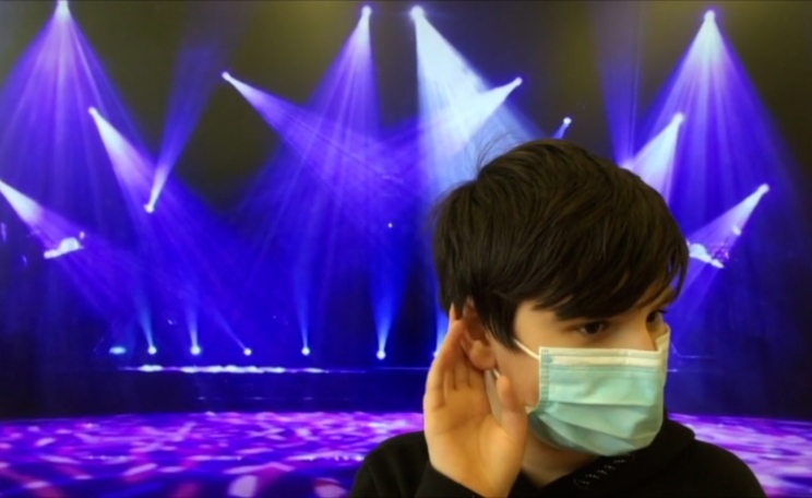 Student wearing a mask cups his hand to his ear like he is trying to listen to something. Behind him is a green-screened image of a concert stage with purple and blue lights.