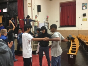 Three middle school students are holding hands and standing in a circle in a school auditorium.
