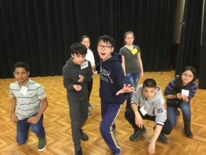 Seven middle school students on an auditorium stage are all leaning toward the camera. Two are lunging. Two are crouching. One is kneeling.