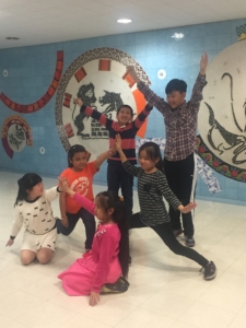 Six students in a school hallway with a colorful mural behind them. They are creating a group pose. All have their arms extended. Two are kneeling. Two are lunging. Two are standing up straight.