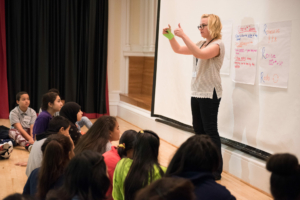 A teaching artist stands in front of a class of students seated on an auditorium stage. The artist shows two thumbs up to the class.