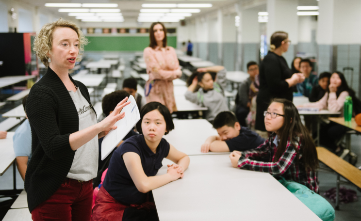 A teaching artist speaks to a class seated at cafeteria tables and gestures with her hands.