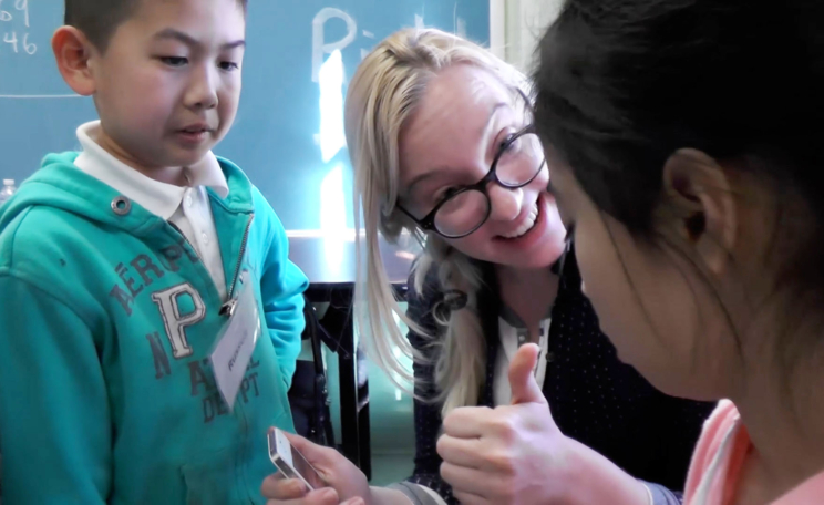 A teaching artist smiles and give the thumbs up sign to a student while a second student looks on.