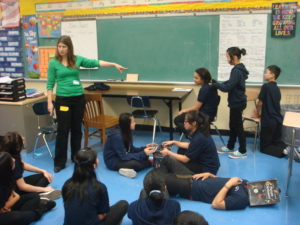 In a classroom, a teaching artist is pointing with her left hand at three students in a theater tableau. The three students are in a line: one is seated, the next is standing behind her holding her ponytail and the third is kneeling behind. In the foreground, two students are seated cross-legged facing each other and stacking books and one student is laying on her back with a book covering her face. Additional students are seated as an audience and looking at the tableau.