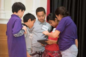 Five middle school students, standing in a tight group, looking at and gesturing towards an iPad.