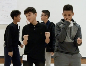 Four middle students are in a scene on the stage of a school auditorium. One of the students in the front is pantomining holding a steering wheel. The student next to him holds his hand near his mouth as if speaking into a handheld radio.