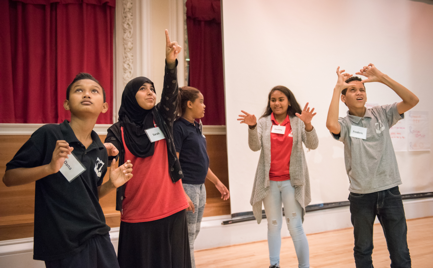 Five middle school students stand on a stage. One is gesturing with her hands as if she is directing the others. The other students are in a tableau, all looking up.