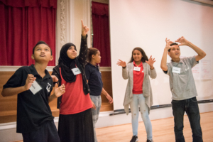 Five middle school students stand on a stage. One is gesturing with her hands as if she is directing the others. The other students are in a tableau, all looking up.