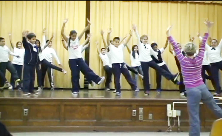 a group of students in white shirts and black pants all dance with their arms stretched up and balancing on their right leg