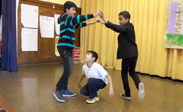 Three elementary school students dance together on the stage. Two students clasp hands and rotate around a student in the center who is in a low pose.