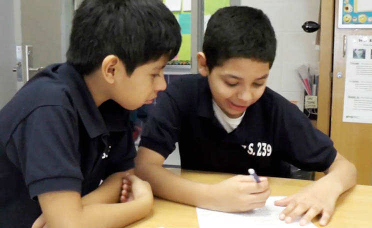 Two elementary school students are sitting at a desk working together. One student writes on a piece of paper.