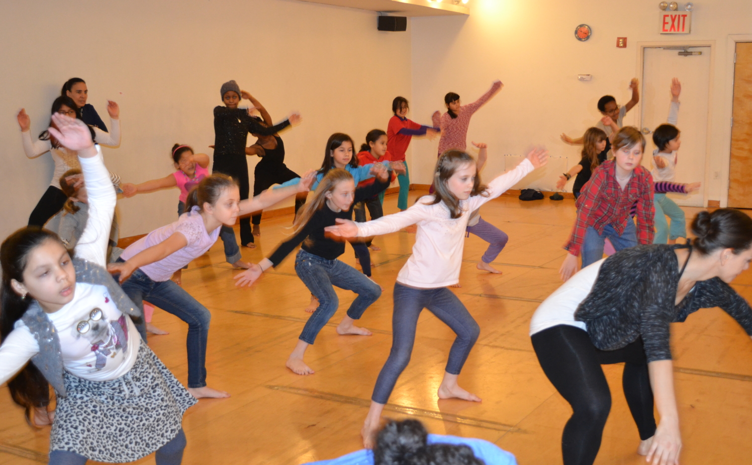a class full of young dancers in their street clothes. Arms are stretched wide. All students are facing the front.