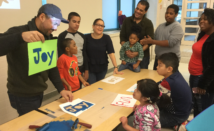 Artist-holds-up-image-of-joy-parents-and-kids-surround-a-table-with-other-prints-of-images-on-it
