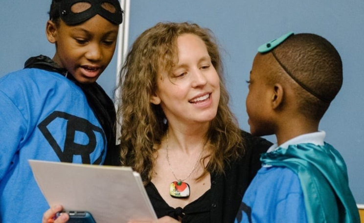 White woman with wavy hair holds a card while two small children look at it with her.