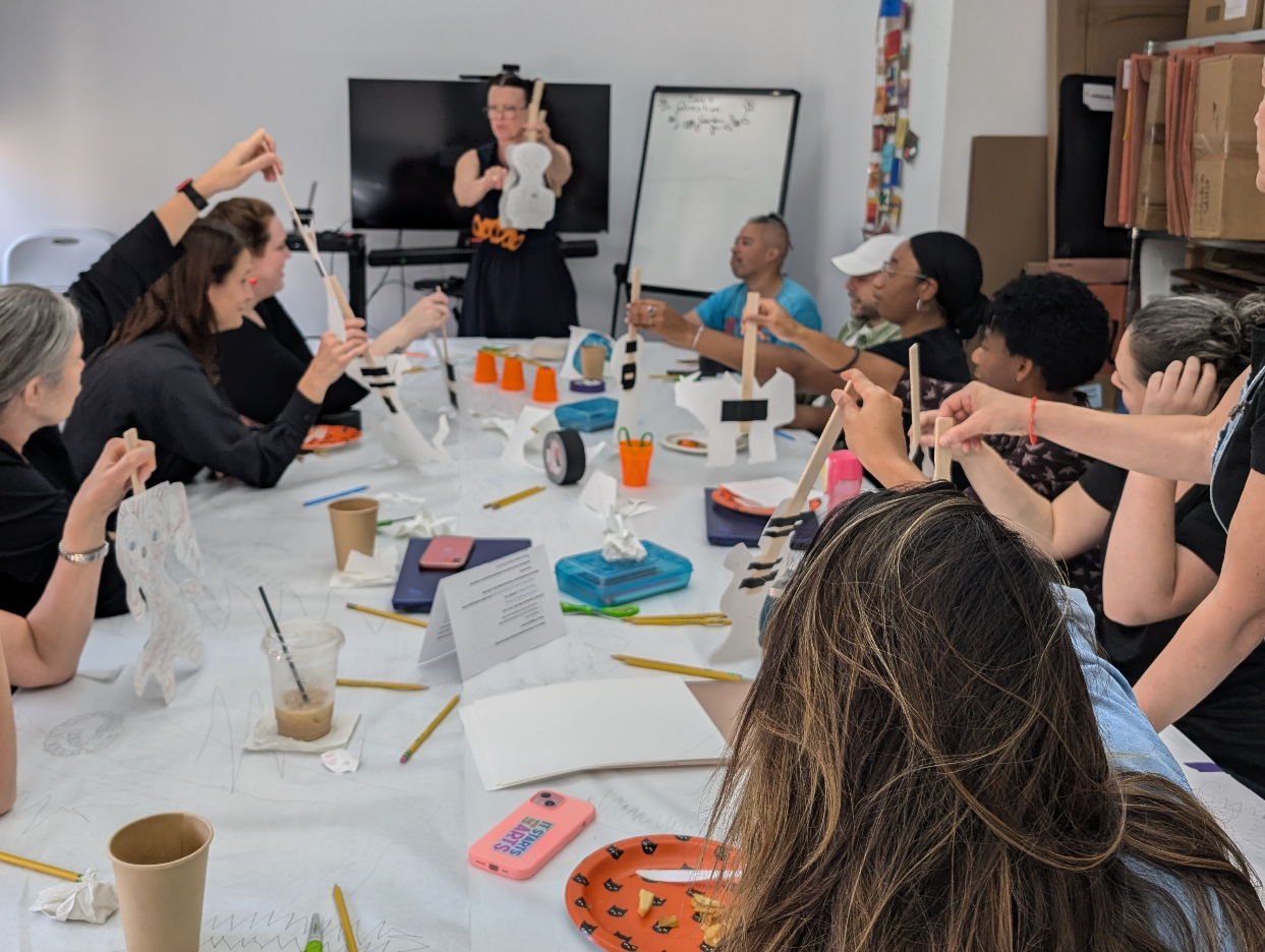 Teacher stands at front of room holding up a puppet on a stick, around a table filled with handouts, coffee, and art supplies. Other adults sit also holding up puppets.