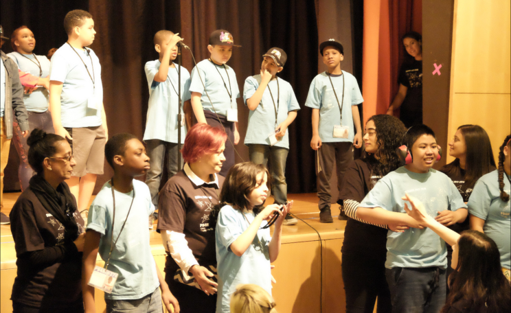 Students are on stage, and in front of the stage. Behind the front row of students are paraprofessionals supporting individual students.