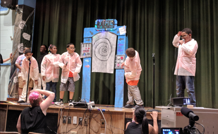Students are performing. In the middle of the stage is a giant cardboard time machine. In front of the stage are two teachers supporting