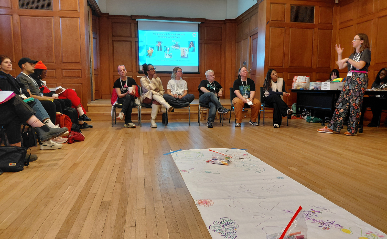 a group of artists sit in front of a slide reading panel. in the forefront of the image is a large piece of butcher block paper that has drawings on it. On the right side a women in flower pants in animatedly introducing the group