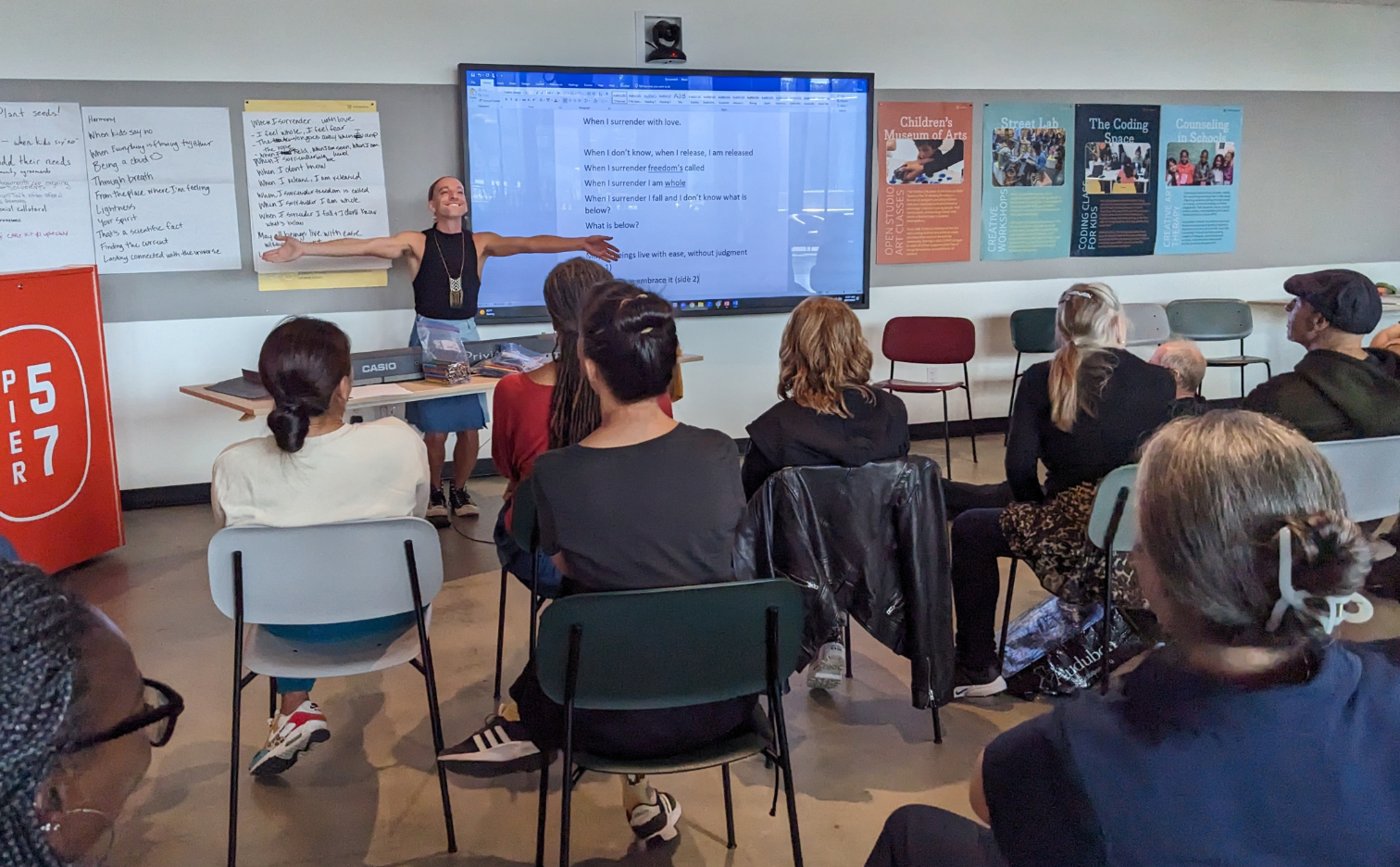 an artist stands at the front of the room - joy on their face arms spread wide. Participants sit facing them, there are lots of filled chart papers on the wall.